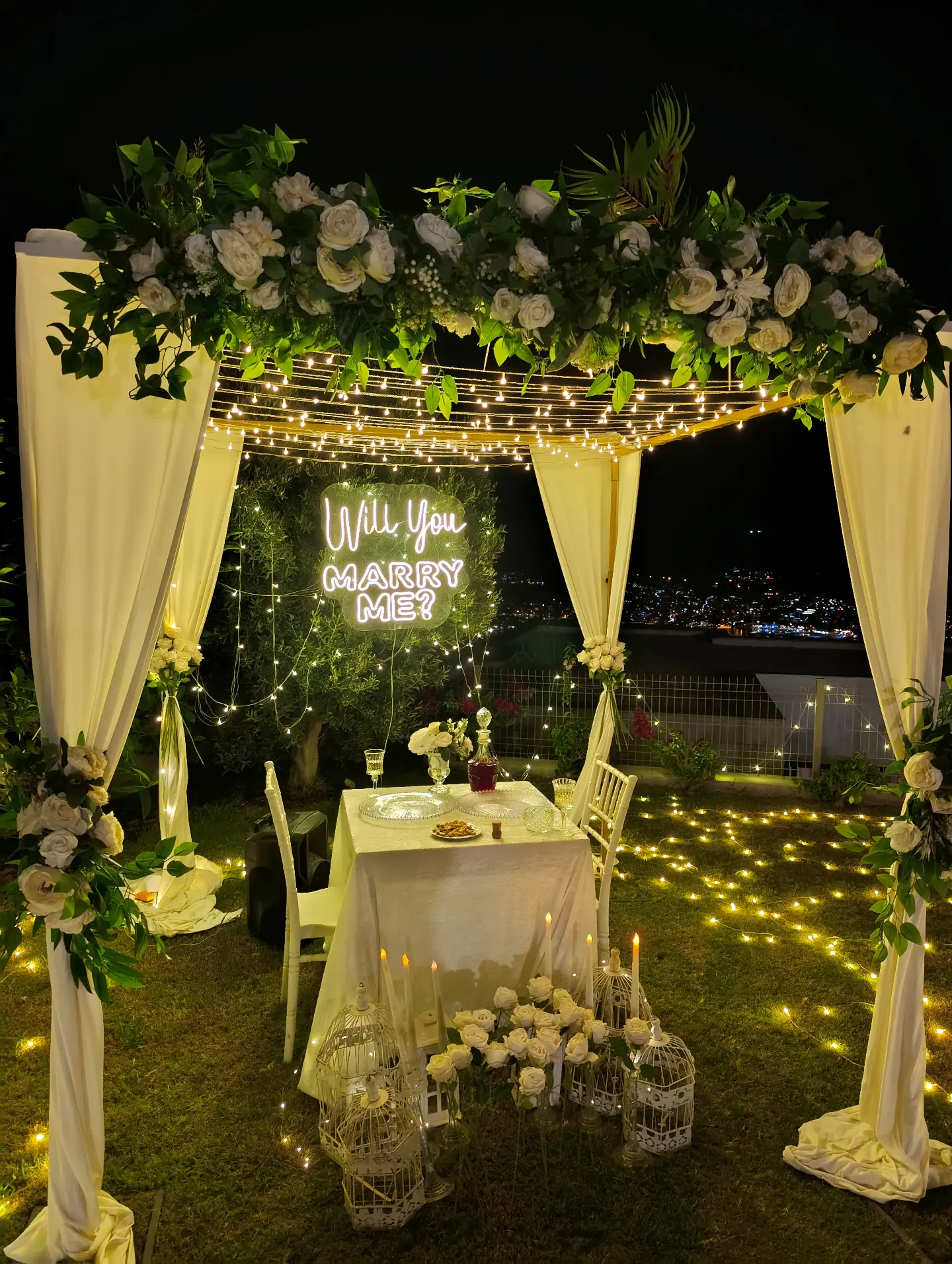Proposal setup with white drapes, floral arch and Will You Marry Me sign
